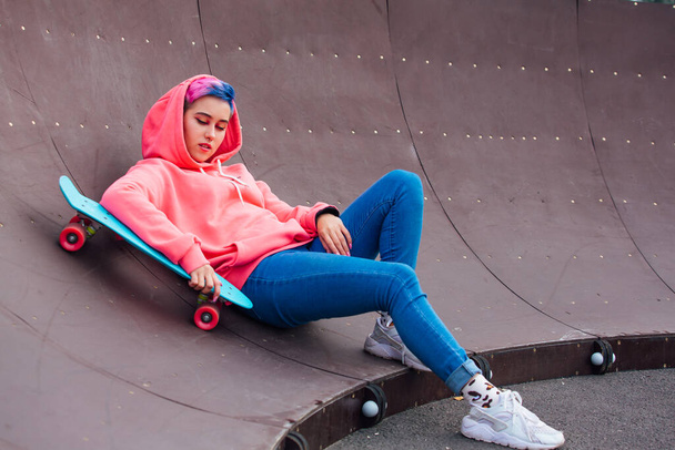 Stylish young woman with short colored hair laying with her blue plastic skateboard in skatepark. Youth concept. - Foto, imagen