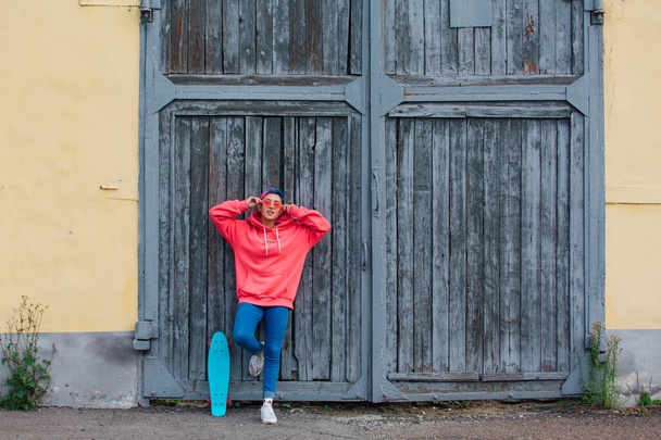 Stylish young woman with short colored hair, dressed in a pink hoodie and pink glasses standing near old wooden gates with her plastic skateboard. Youth concept. - Photo, Image