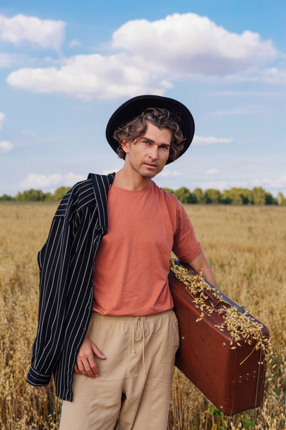 Rural Countryside Scene. Tall handsome man dressed in a black shirt and black hat holding brown vintage leather suitcase full of ears of oats at golden oat field - Photo, image