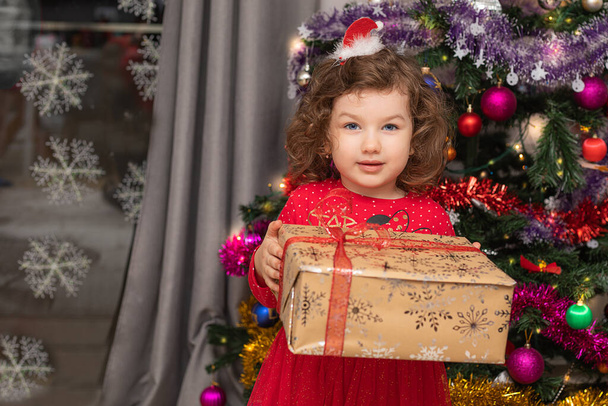 Veselé Vánoce. Veselá krásná holčička sedící u vánočního stromku a držící dárkovou krabici. Vánoční zázraky. Šťastný Nový rok. Domácí život - Fotografie, Obrázek