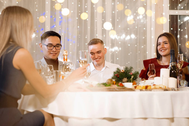 Egy csoport többnemzetiségű ember ül az ünnepi asztalnál egy pohár pezsgővel, mosolyog, beszél egy étteremben. Szelektív fókusz, homályos fények. - Fotó, kép