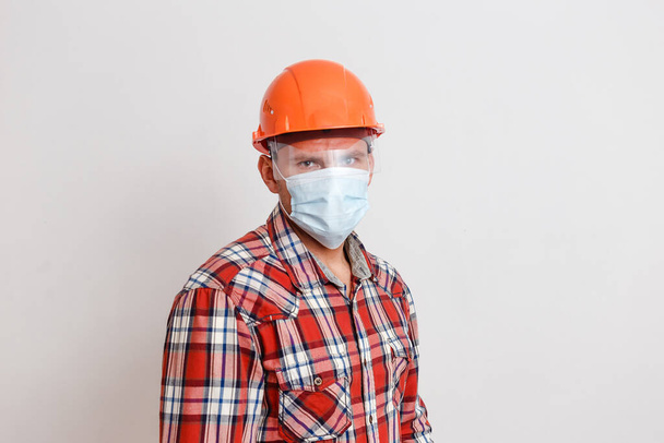a man in a plaid shirt and an orange protective helmet on his head and wearing a medical mask on a white background. mask mode. stop coronavirus - Foto, immagini