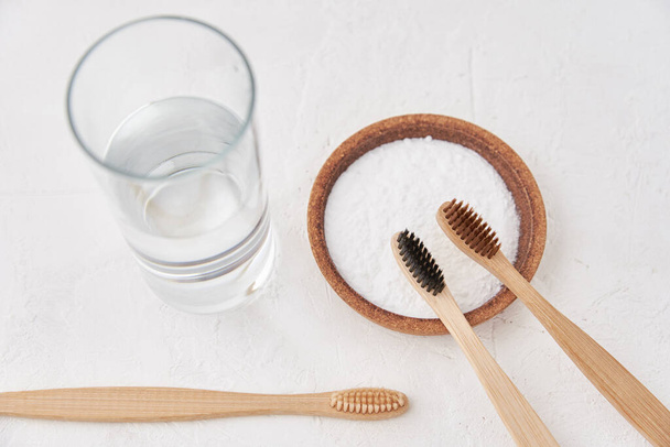 Cepillos de dientes de bambú, bicarbonato de sodio y un vaso de agua sobre un fondo blanco. Cepillos de dientes ecológicos, concepto de cero residuos - Foto, imagen