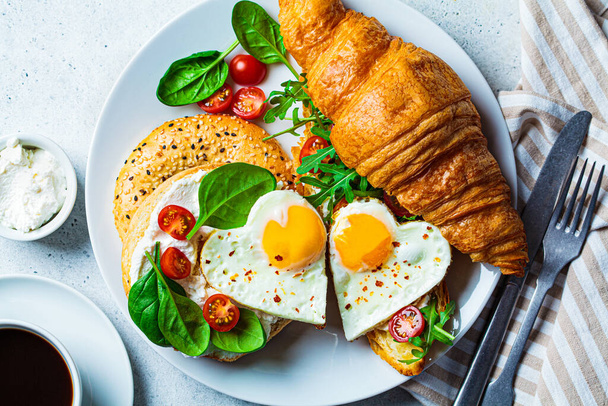 Valentine's day breakfast concept. Sandwich with croissant, bagel, cream cheese and fried eggs hearts on a white plate. - Фото, изображение