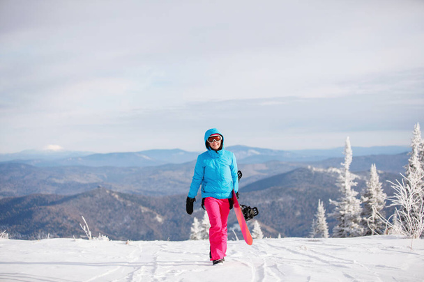 Kadın snowboardcu karla kaplı bir dağda yürüyor. Mavi kapüşonlu ceket, gözlük ve pembe pantolon giyiyor. Sağlıklı bir yaşam tarzı. Spor konsepti. Seçici odak. - Fotoğraf, Görsel