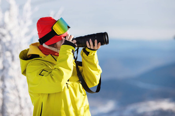 Female photographer in ski equipment takes pictures with a camera with a huge lens. Winter clothes: red pants and a warm yellow jacket, knitted hat, ski goggles. Selective focus. - Φωτογραφία, εικόνα
