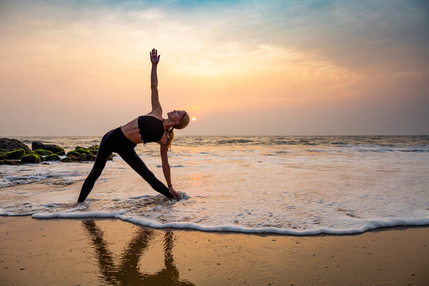 Middelbare leeftijd vrouw in het zwart doet yoga op zandstrand in India staande asana Trikonasana (driehoek pose) bij zonsondergang. Gezonde levensstijl. - Foto, afbeelding
