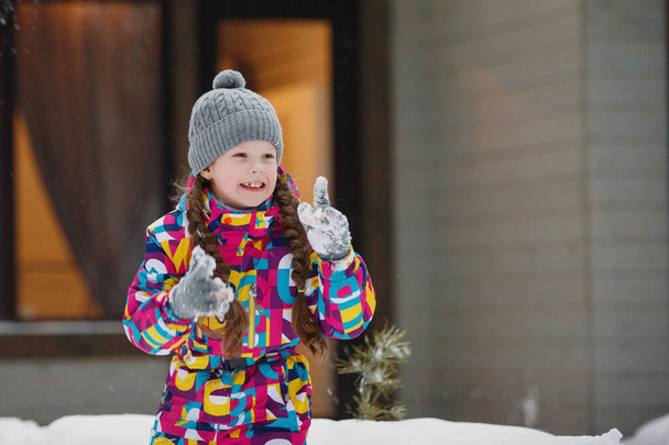 Menina com tranças em uma jaqueta de inverno e um chapéu joga em bolas de neve no alpendre da casa. - Foto, Imagem
