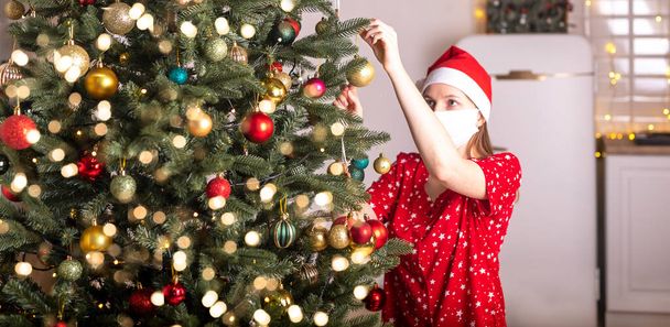 Young woman wearing face mask and pyjamas decorating christmas tree at home alone. celebrating xmas amnd new year during lockdown and pandemic. - Photo, Image
