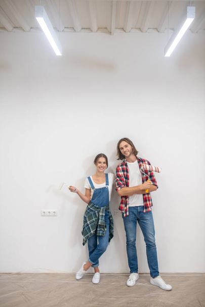 Young couple standing near the wall with paint rollers and hands - Fotó, kép