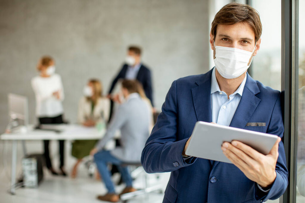 Joven hombre de negocios guapo con máscara facial protectora mientras sostiene la tableta digital en el espacio de oficina - Foto, Imagen