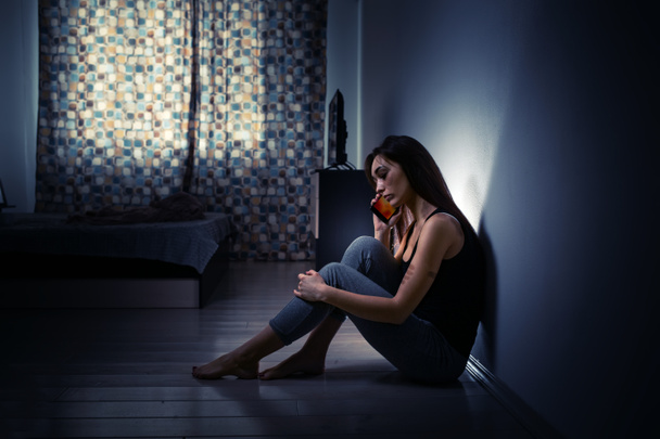 El concepto de violencia doméstica. Mujer caucásica agotada con golpizas en el cuerpo se sienta en el suelo en una habitación oscura, contra la pared y llama a un teléfono móvil. - Foto, Imagen