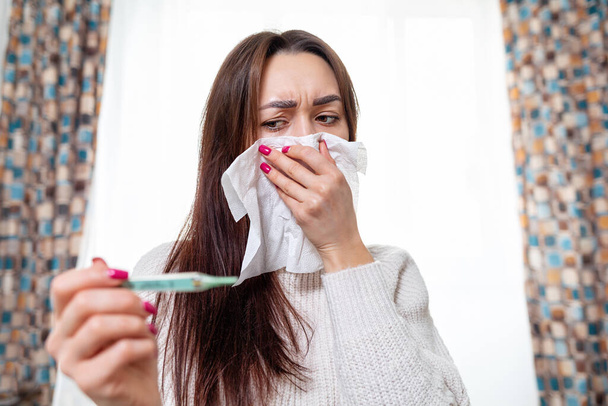 Nuori valkoihoinen nainen näyttää kuumemittarin korkeassa lämpötilassa, peittäen nenänsä nenäliinalla. Hoito kotona. Virusinfektion ja karanteenin käsite. - Valokuva, kuva