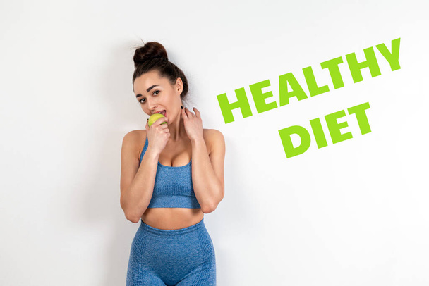 The concept of healthy eating. A young Caucasian woman in sports clothes, with a toned figure, eating a green Apple. White background. The inscription Healthy diet. - Photo, image