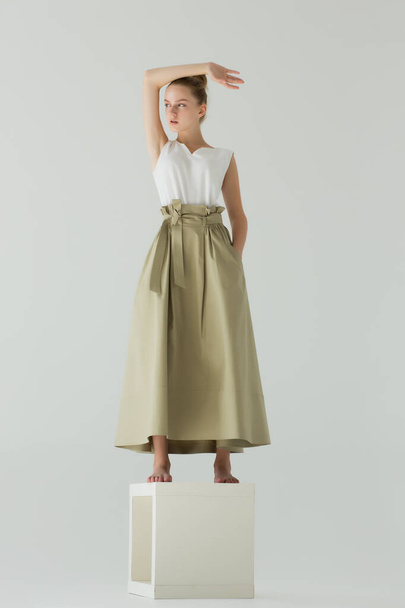 A woman stands in the studio on a white cube. Clothes - a long green skirt and a white T-shirt. The arm is raised up, bent at the elbow. She stands barefoot with one hand in her pocket. - Foto, imagen