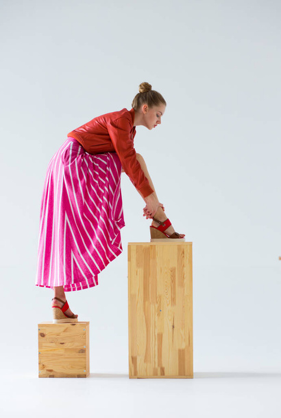 The girl is standing on the cubes. She takes a wide step and bends towards her leg. Clothing - a long skirt with wide stripes, high wedge sandals and an orange jacket. - Fotó, kép