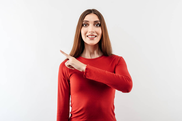 Image of young happy woman with long chestnut hair, wearing in casual red sweater, smiling and pointing forefinger aside, shows place for ad. Studio shot, white background, isolated - 写真・画像