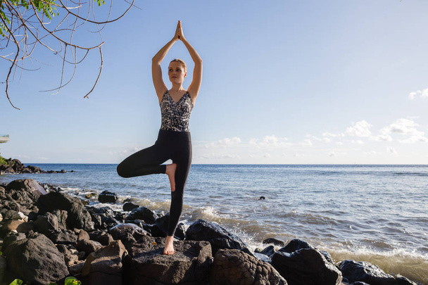 Jeune femme en noir faire du yoga sur la plage de roche pose d'arbre vue sur l'océan - Photo, image