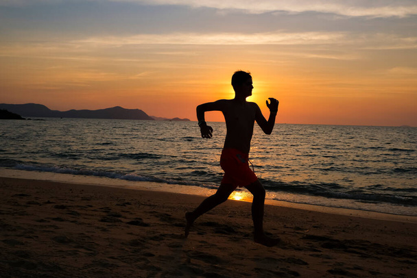 Silhouette of a young man running at the beach at sunrise with the sun in the background. Running on the sea shore in the evening. Empty beach. Staying fit and healthy. Copy space banner. - Φωτογραφία, εικόνα