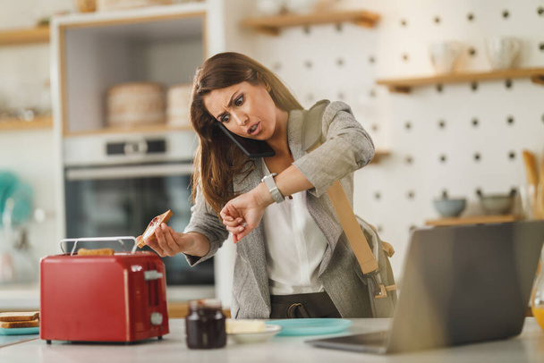 Foto de una joven mujer de negocios con múltiples tareas desayunando y mirando en su cocina mientras se prepara para ir a trabajar. - Foto, Imagen