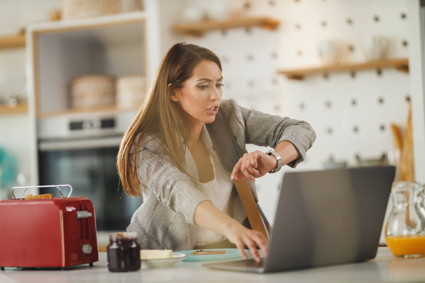Ammuttu monitoiminen nuori liiketoiminnan nainen katselee katsella ja käyttää kannettavaa tietokonetta hänen keittiössä samalla saada valmis menemään töihin. - Valokuva, kuva