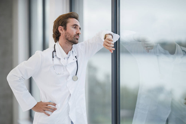 Un médico cansado parado cerca de una ventana y mirando el reloj en un pasillo vacío del hospital durante la pandemia de Covid-19. - Foto, Imagen