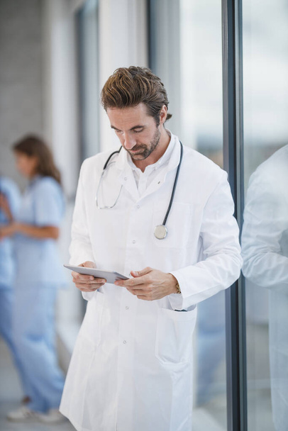 Inyección de un médico maduro usando tableta digital mientras estaba de pie cerca de una ventana en el pasillo de un hospital durante la pandemia de Covid-19. - Foto, imagen