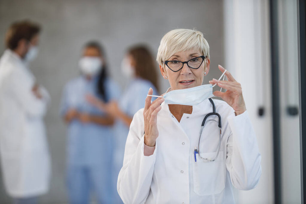 Een vermoeide senior arts die een beschermend masker vasthoudt tijdens een korte pauze in een ziekenhuisgang tijdens de Covid-19 pandemie. Kijkend naar camera. - Foto, afbeelding