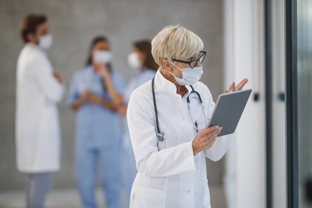 Snímek starší doktorky s ochrannou maskou, která během pandemie Covid-19 volala na digitální tablet v nemocniční chodbě. - Fotografie, Obrázek
