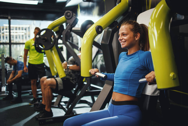 Una foto de una joven musculosa en ropa deportiva haciendo ejercicio en un entrenamiento cruzado en el gimnasio. Ella está haciendo ejercicio para su pecho en una máquina. - Foto, imagen