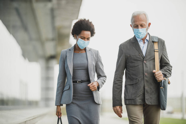 Снимок успешной черной бизнесвумен и ее старший коллега мужчина в защитной маске и говорить во время прогулки перед офисным зданием во время пандемии COVID-19. - Фото, изображение