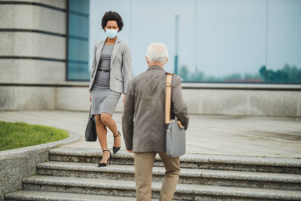 Aufnahme zweier erfolgreicher multiethnischer Geschäftsleute mit Schutzmaske beim Treppensteigen vor dem Bürogebäude während der COVID-19-Pandemie. - Foto, Bild