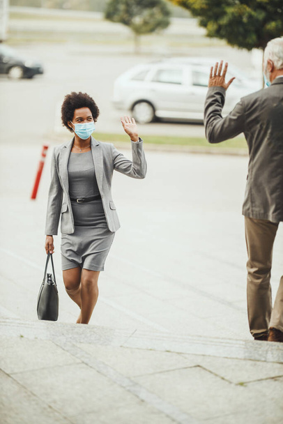 Shot of successful black businesswoman and her senior male colleague using protective mask and greeting with wave hands during COVID-19 pandemic to avoid handshakes. - Photo, Image