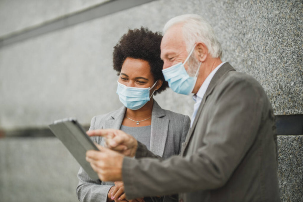 Shot of successful black businesswoman and her senior male colleague with protective mask using digital tablet and having a discussion while standing against a wall of corporate building during COVID-19 pandemic. - Photo, Image