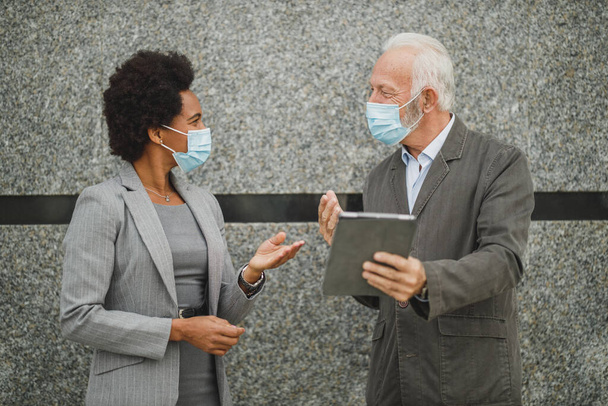 Aufnahme zweier erfolgreicher multiethnischer Geschäftsleute mit Schutzmaske, die ein digitales Tablet benutzen und eine Diskussion führen, während sie während der COVID-19-Pandemie vor einer Wand eines Firmengebäudes stehen. - Foto, Bild