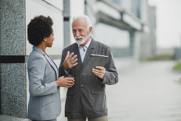 Snímek úspěšného staršího podnikatele a jeho černošky kolegyně mluvit a pomocí digitálního tabletu během přestávky na kávu před kancelářskou budovou. - Fotografie, Obrázek