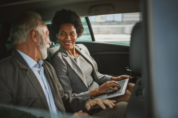 Prise de vue de deux personnes multi-ethniques utilisant un ordinateur portable et parlant de leur travail assis sur le siège arrière d'une voiture pendant leur trajet professionnel. - Photo, image
