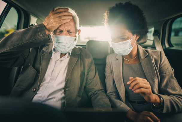 Disparo de dos personas multiétnicas preocupadas con máscara protectora usando el portátil y teniendo una discusión mientras están sentados en el asiento trasero de un automóvil en su viaje de negocios durante la pandemia de COVID-19. - Foto, Imagen