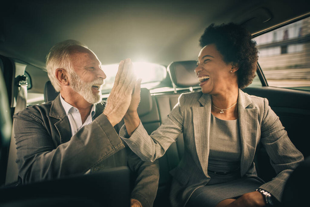 Shot van twee succesvolle multi-etnische mensen geven vijf en vieren goed werk terwijl zitten op de achterbank van een auto tijdens hun zakelijke woon-werkverkeer. - Foto, afbeelding