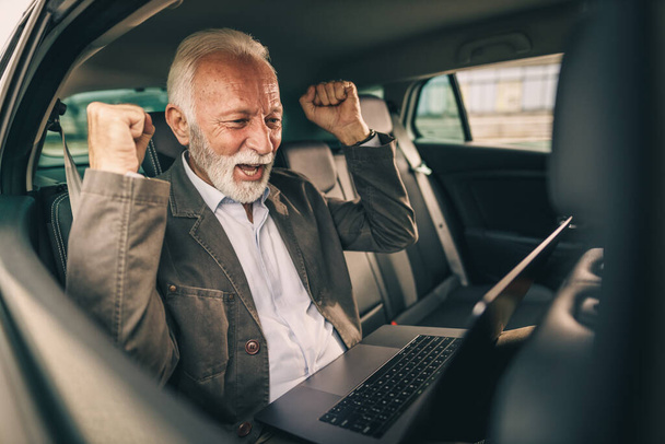 Πυροβολισμός ενός χαρούμενου ηλικιωμένου άνδρα που χρησιμοποιεί φορητό υπολογιστή και γιορτάζει την επιτυχία του ενώ κάθεται στο πίσω κάθισμα ενός αυτοκινήτου κατά τη διάρκεια της επαγγελματικής του μετακίνησης. - Φωτογραφία, εικόνα