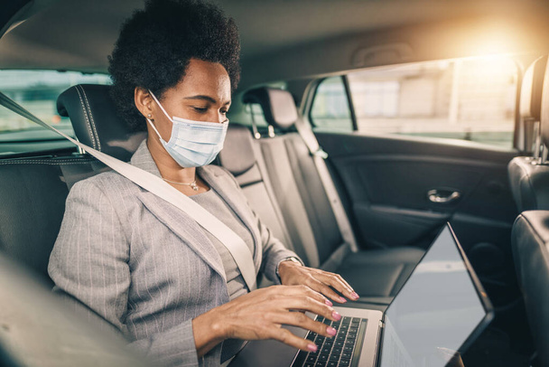 Opname van een succesvolle zwarte vrouw met beschermend masker werkend op laptop terwijl ze op de achterbank van een auto zat te pendelen tijdens COVID-19 pandemie. - Foto, afbeelding