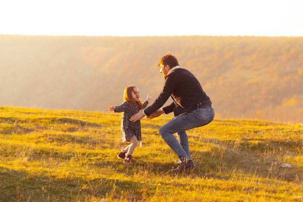 Family vacantion. Photo of father catching his daughter, meadow or park on sunset - 写真・画像