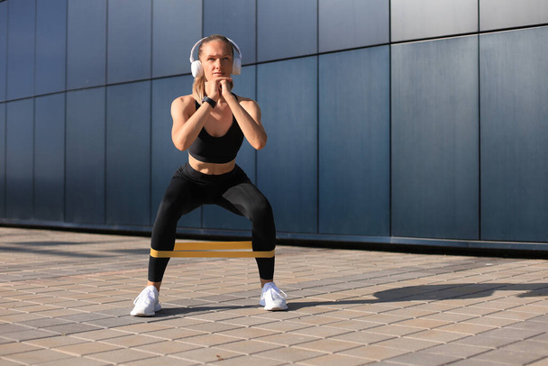 Sporty woman doing squats with fitness gum expander outdoors - Fotoğraf, Görsel