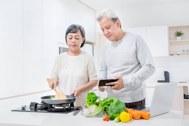 Senior couple cooking together in the kitchen while looking recipe on a smartphone. Shot at home - 写真・画像