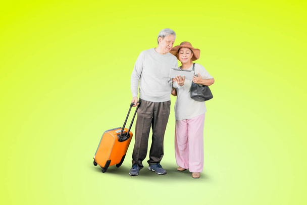 Senior couple using a digital tablet while carrying a luggage for vacation and standing in the studio with green screen background - Fotó, kép