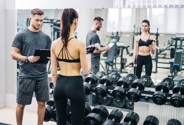 Beautiful young woman with her personal trainer at the gym discuss the progress on a clipboard held by the man. Healthy lifestyle. - Фото, изображение
