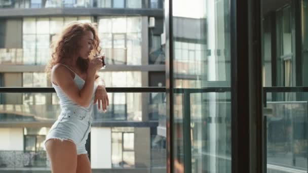 A young woman is standing on the balcony and drinking coffee. She is enjoying the weather. 4K - Footage, Video