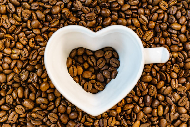 Coffee time. White cup in shape of heart on roasted coffee beans background. Top view - Photo, Image