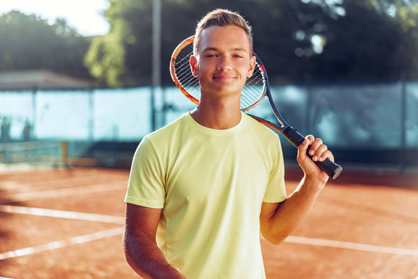 Young man teenager with tennis racket standing near net on clay court - Фото, зображення