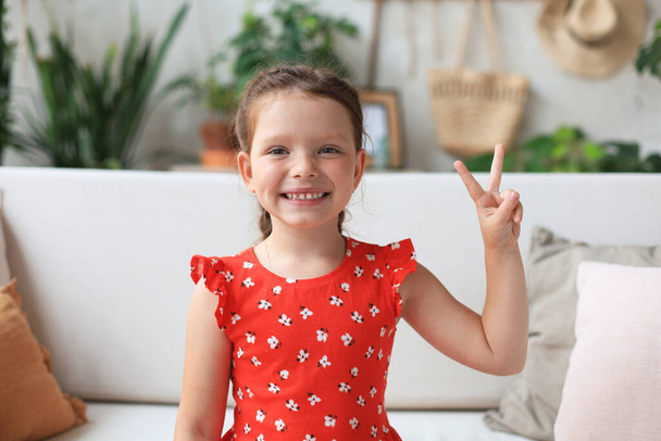 Kis kaukázusi lány piros ruhában ül a kanapén, mosolyogva boldog arccal győzelmi jelet csinál. - Fotó, kép